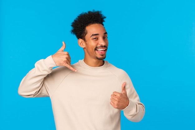 Appelez-moi plus tard bébé. Homme macho confiant afro-américain impertinent et coquet avec moustache, coupe de cheveux afro montrant le pouce vers le haut et le geste du téléphone, clin d'œil et sourire affectueux, mur bleu