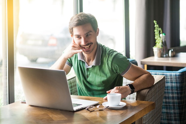 Appelle-moi. Jeune homme d'affaires drôle en t-shirt vert assis, regardant l'écran d'un ordinateur portable lors d'un appel vidéo et demandant à le rappeler bientôt. entreprise, concept indépendant. tir à l'intérieur près de la fenêtre pendant la journée.