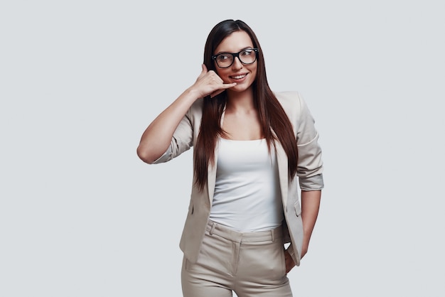 Appelle moi! Jolie jeune femme gesticulant et souriant en se tenant debout sur fond gris