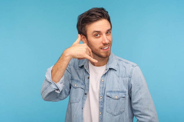 Appelle-moi. Portrait d'un bel homme joyeux en chemise en jean debout avec un geste de la main téléphonique et souriant à la caméra, flirtant en proposant de contacter par téléphone. tourné en studio isolé sur fond bleu