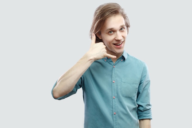 Appelle-moi. Portrait de drôle beau jeune homme blond aux cheveux longs en chemise décontractée bleue debout avec la main de geste d'appel et regardant la caméra. tourné en studio intérieur, isolé sur fond gris clair.