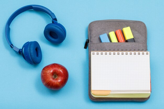 Apple, écouteurs et cahier d'exercices ouvert sur étui à crayons avec feutres de couleur et marqueur sur fond bleu. Vue de dessus. Retour au concept de l'école. Fournitures scolaires.