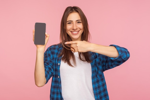 Application, publicité sur appareil mobile. Heureuse fille souriante en chemise à carreaux pointant vers l'écran du téléphone portable, montrant l'affichage, maquette pour le texte commercial. tourné en studio isolé sur fond rose, espace de copie