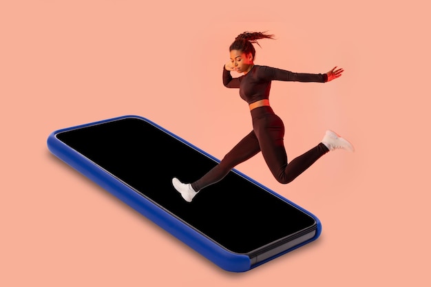 Application de remise en forme Femme noire sautant sur l'écran vide du smartphone ayant une séance d'entraînement sur la maquette de fond de studio de pêche