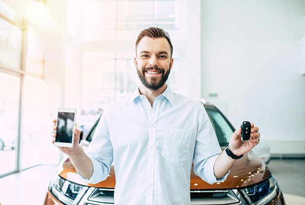 Photo applications pour acheter une voiture facilement un jeune homme barbu montre une clé de voiture et un téléphone avec un écran vide sur l'appareil photo d'un concessionnaire