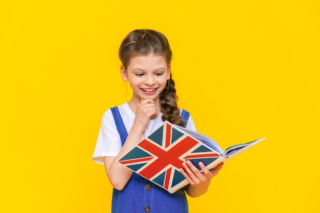 Apprendre l'anglais pour les enfants Une petite fille tient un livre avec un drapeau anglais dans ses mains et sourit largement sur un fond isolé jaune