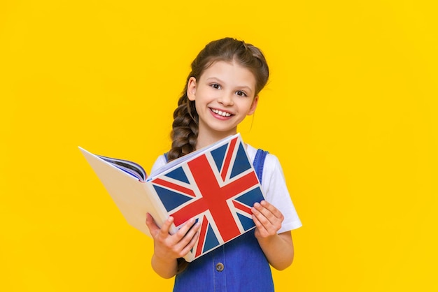 Apprendre l'anglais pour les enfants Une petite fille tient un livre avec un drapeau anglais dans ses mains et sourit largement sur un fond isolé jaune