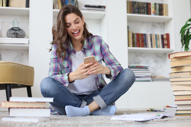 Apprentissage à distance. Jeune étudiante vérifiant les médias sociaux avant de reprendre ses études, assise sur le sol contre un intérieur domestique confortable, entourée d'une pile de livres