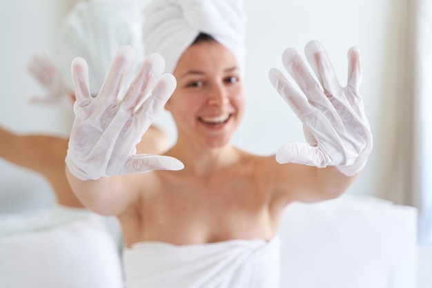 Après une douche, une fille enveloppée dans une serviette utilise des gants cosmétiques pour hydrater la peau de ses mains