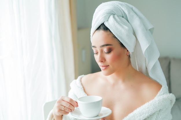 Après une douche, une jeune femme en peignoir avec une serviette sur la tête boit du café et regarde par la fenêtre