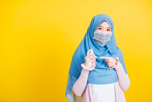 Arabe musulman asiatique, Portrait d'une belle jeune femme heureuse Islam religieux porter voile hijab et masque facial protéger elle met en quarantaine la maladie coronavirus pointer l'alcool vaporiser sur les mains isolé jaune backgr