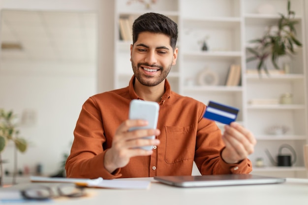 Un arabe souriant utilisant un téléphone et une carte de crédit à la maison