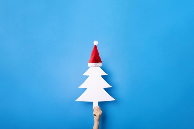 Arbre de Noël blanc en chapeau de Noël dans les mains sur fond bleu, concept de joyeux Noël