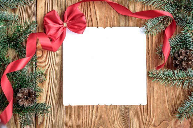 Arc rouge décoratif, ruban et enveloppe avec note de carte blanche pour le texte. Composition de Noël avec des branches de sapin, des cônes et des boules sur planche de bois. Copiez l'espace.