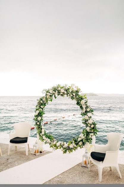 L'arche de mariage ronde faite de roses et de feuilles vertes se dresse sur la jetée entre deux fauteuils