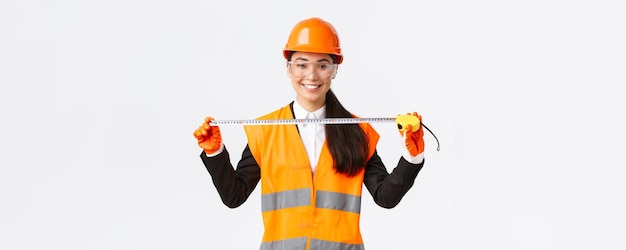 Architecte asiatique professionnelle confiante mesurant la disposition portant un casque de sécurité et un uniforme et tenant un ruban à mesurer souriant satisfait satisfait du résultat obtenu pendant la construction