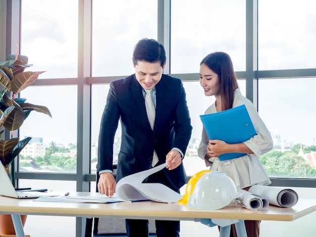 Architecte en chef masculin asiatique ou ingénieur en costume et jeune femme secrétaire discutent du plan avec un ordinateur portable et des casques blancs et jaunes sur un bureau sur une immense vitre au bureau.