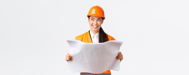 Architecte féminine asiatique créative imaginant son plan debout dans un casque de sécurité et regardant loin pensivement tout en lisant des plans illustrant la construction après la fin des travaux de construction fond blanc