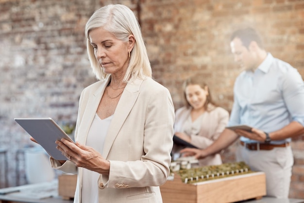 Architecte mature utilisant une tablette pour analyser les conceptions dans un bureau moderne Femme d'affaires senior avec un arrière-plan flou d'une jeune équipe d'architectes travaillant ensemble sur un modèle de bâtiment