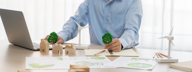 L'architecte professionnel prévoit d'utiliser la conception verte dans une maison écologique. L'ingénieur qualifié conçoit un plan de maison écologique en utilisant un ordinateur portable sur la table avec la maison et le modèle de moulin à vent dispersés autour de la délimitation.