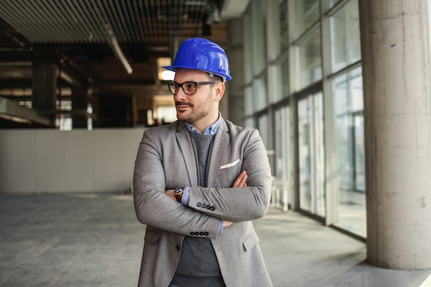 Architecte souriant debout dans le bâtiment en processus de construction avec les bras croisés