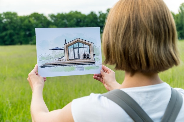 Architecte tenant un croquis dessiné à la main d'une maison de grange devant une parcelle de terrain