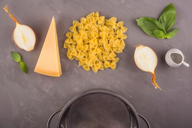 Photo les arcs de pâtes farfalle crues sont disposés exactement sur un fond gris avec du basilic et une casserole pour la cuisson