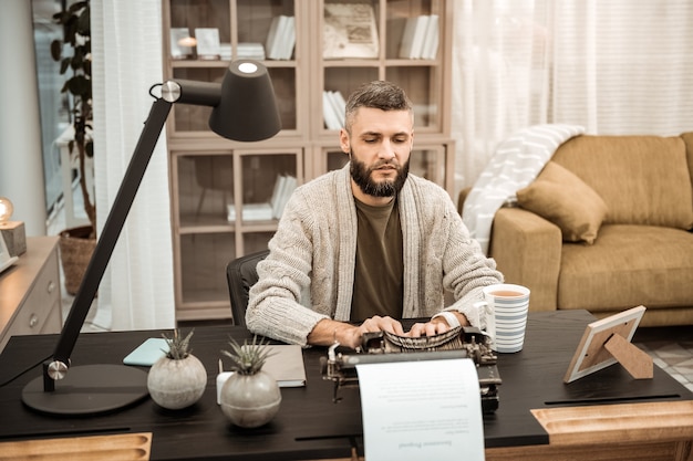 Armoire bien équipée. Homme concentré sérieux en cardigan gris tapant sur une machine à écrire tout en passant du temps dans une armoire à la mode