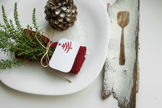 Arranger la table pour le concept des vacances d'hiver. Décoration de table dans un style minimaliste, serviette rouge aux herbes, contour d'une cuillère à dessert, pomme de pin et carte de place de vacances