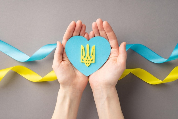 Arrêtez le concept de guerre en Ukraine Photo vue de dessus à la première personne de mains féminines tenant un coeur bleu avec un trident doré sur le drapeau national sur les paumes sur fond gris