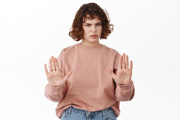 Arrêtez, reculez. Fille bouclée sérieuse, montrant un geste d'interdiction de bloc, rejetant quelque chose, signe de rester à l'écart, disant à la distance sociale, debout sur fond blanc