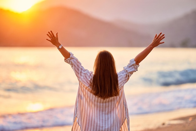 L'arrière d'une femme libre heureuse et sereine calme à bras ouverts profite d'un beau moment de vie au bord de la mer au coucher du soleil