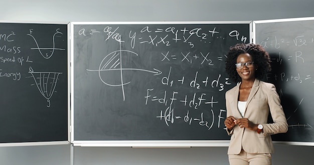 L'arrière de la jeune femme afro-américaine professeur de mathématiques ou de physique écrit des formules sur tableau noir à la craie. Portrait de femme joyeuse tuteur se tournant vers la caméra et souriant joyeusement. Vue arrière.