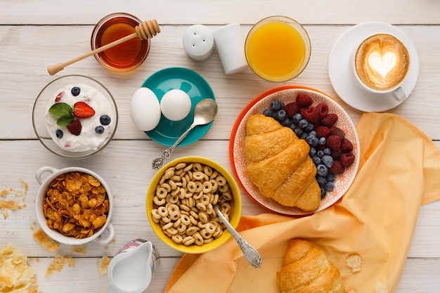 Arrière-plan du menu du petit-déjeuner riche. Croissants croustillants français, muesli, verre d'orange fraîche, yaourt, café, baies, miel et œufs durs pour de savoureux repas du matin sur une table en bois, vue de dessus