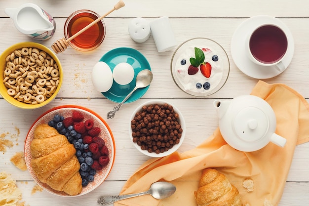 Arrière-plan du menu du petit-déjeuner riche. Croissants croustillants français, muesli, yaourt, café, baies, miel et œufs durs pour de savoureux repas du matin sur table en bois, vue de dessus