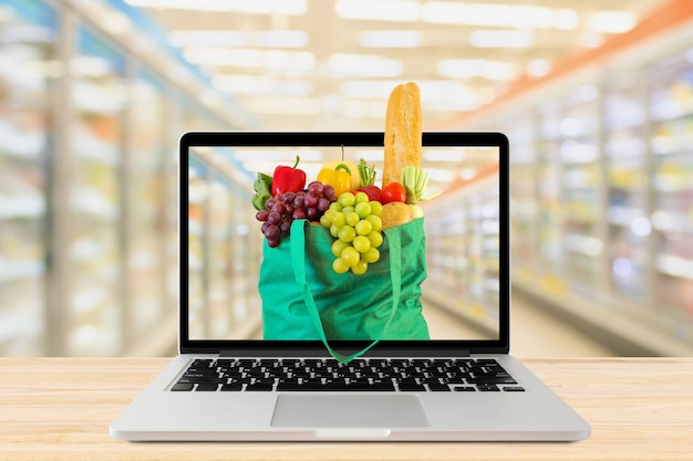 Arrière-plan flou allée de supermarché avec ordinateur portable et sac à provisions vert sur table en bois épicerie en ligne concept