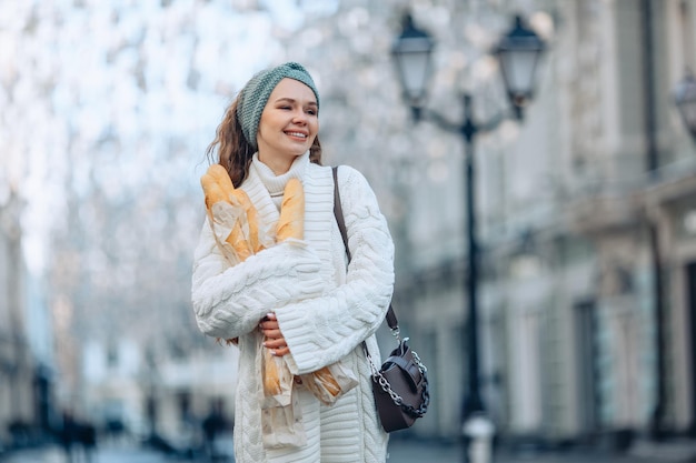 Arrière-plan flou de portrait de rue élégant urbain. Une jeune femme impressionnante vêtue d'un costume doux en tricot blanc et d'une bande tricotée bleue tient plusieurs paquets dans ses mains. Retour de la boulangerie.