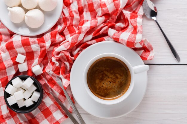 Arrière-plan de l'heure du café. Friandises et tasse de café sur un tissu à carreaux blancs et rouges. Sucre en morceaux et meringues sur bois blanc, vue de dessus, espace de copie