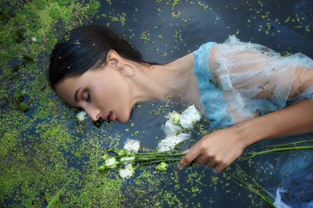 Art belle femme romantique se trouve dans les marais en robe longue bleue avec des fleurs. Portrait brune en robe transparente dans les lentilles d'eau de boue de marais d'eau. Couverture du livre