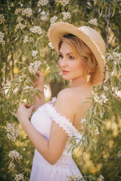 Art mode portrait d'une belle jeune femme blonde dans un jardin fleuri en été vert dans une robe blanche légère, dans un chapeau de paille. Fille à la campagne