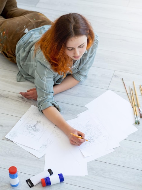 Art de la peinture Passe-temps créatif Loisirs à la maison relaxants Artiste féminine inspirée appréciant le dessin de croquis au crayon sur papier allongé sur le sol avec des pinceaux de peinture en studio léger