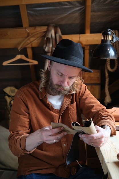 Artisan barbu au chapeau assis sur son lieu de travail et examinant des échantillons de cuir avant de fabriquer des chaussures en atelier