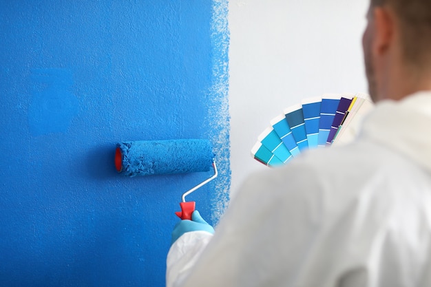 L'artisan tient le rouleau et une palette de couleurs et peint le mur blanc en bleu. Services de peinture murale et concept de peinture