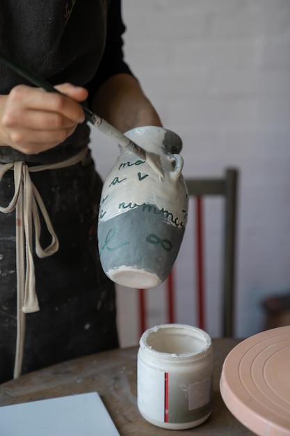 Une artisane en tablier crée de la vaisselle en céramique peignant un vase blanc avec un pinceau en atelier