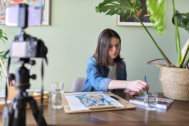 Artiste, adolescente, dessine et enregistre sur caméra vidéo pour son blog. Peintures, dessins sur une table à la maison, appareil photo sur trépied, dessin racontant la blogueuse vlogeuse. Technologie, art, jeunesse, concept d'éducation