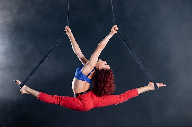 Artiste féminine de cirque aérien athlétique, sexy et flexible avec rousse sur des sangles aériennes sur fond noir.