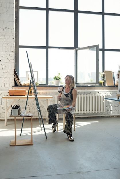 Artiste féminine détendue en tablier tenant la palette et boire du vin tout en pensant à la peinture