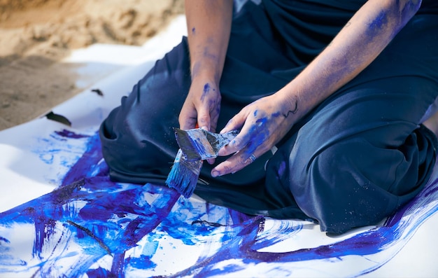 Artiste féminine en robe bleu foncé enduite de peinture à la gouache bleue avec de larges traits sur une grande toile allongée sur la plage, gros plan. Performance expressive, art contemporain, plein air