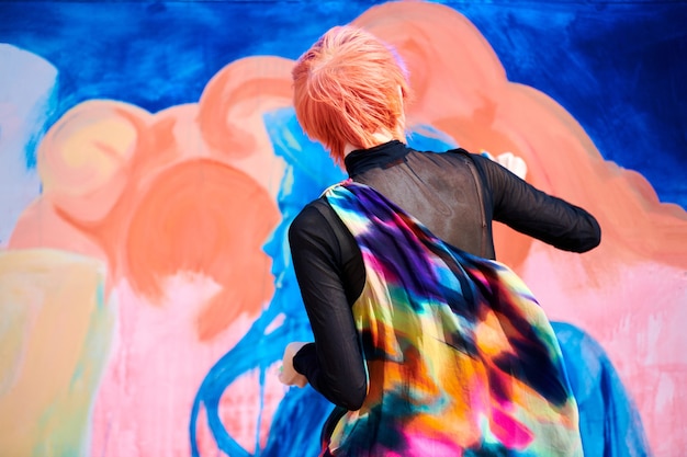 Artiste de la jeune femme aux cheveux courts multicolores dans des vêtements colorés lumineux peignant une image abstraite tenant un pinceau large. Peinture à l'huile sur grande toile, fragment d'œuvre d'art de couleur orange, bleu, rose