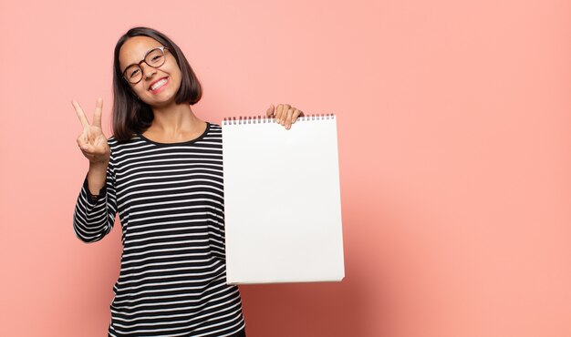 Artiste de la jeune femme souriante et à la recherche heureuse, insouciante et positive, gesticulant la victoire ou la paix d'une main